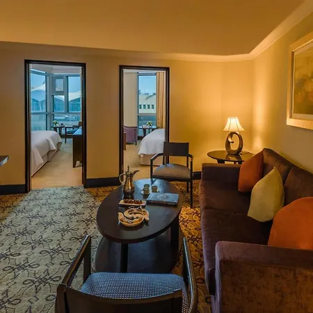 Pullman Zamzam Makkah Hotel Mecca Exterior photo The photo shows a well-furnished hotel suite. In the foreground, there's a cozy seating area with a sofa adorned with colorful cushions, a coffee table, and a small decorative item on it. A lamp provides a warm glow. In the background, two doorways l
